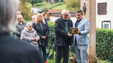 Vor einer Stele zwei Männer. Sie schauen auf einen Bilderrahmen. Links daveon einige Teilnehmende der Veranstaltung.
