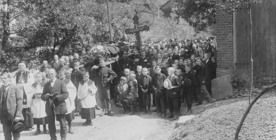 Historische Schwarz-weiß Aufnahme. Eine große Gruppe von Menschen jeden Alters in Festtagskleidung umrahmen einen Sarg. Hinter dem Sarg wird ein großes Kreuz getragen.