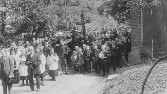 Historische Schwarz-weiß Aufnahme. Eine große Gruppe von Menschen jeden Alters in Festtagskleidung umrahmen einen Sarg. Hinter dem Sarg wird ein großes Kreuz getragen.