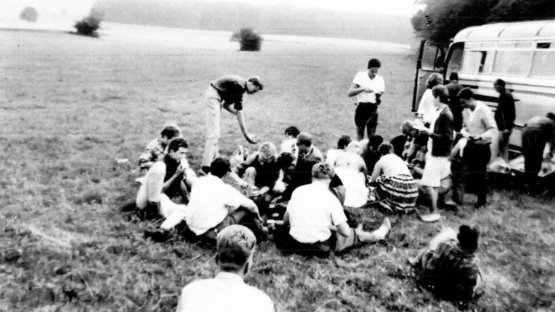 Historisches chwarz-weiß Foto. Eine Gruppe von Jugendlichen sitzt am Rand eines Feldes. Rechts dahinter eine Bus.