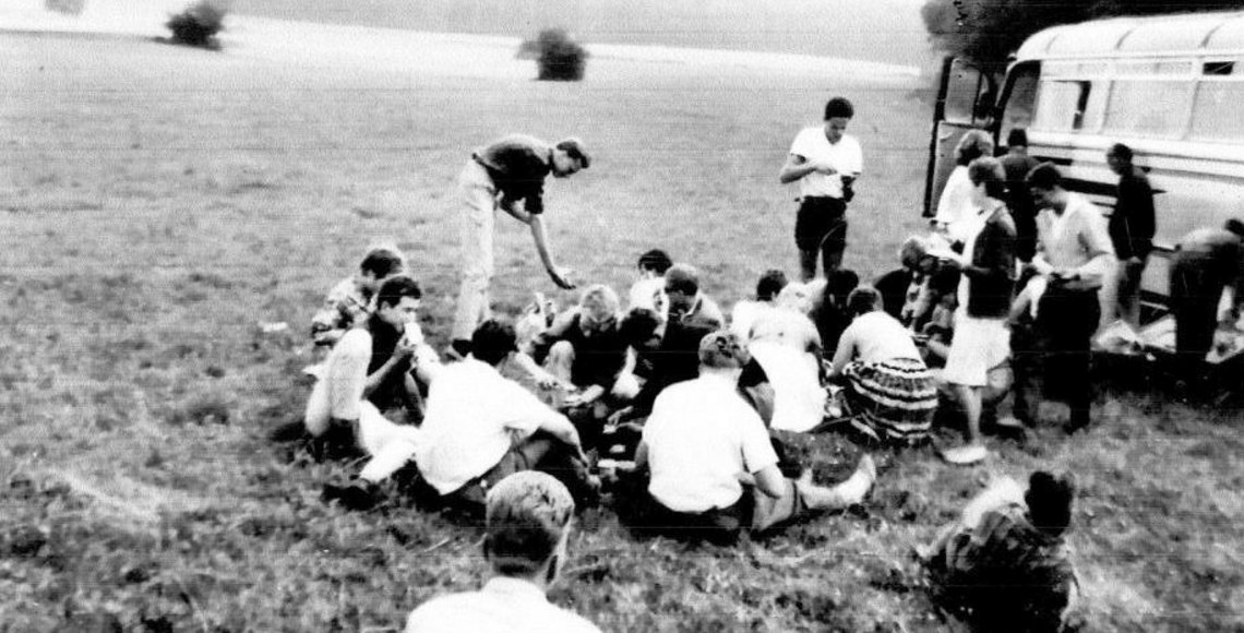 Historisches chwarz-weiß Foto. Eine Gruppe von Jugendlichen sitzt am Rand eines Feldes. Rechts dahinter eine Bus.