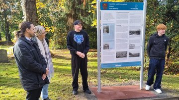 Zwei Schüler stehen links und rechts der Tafel. Links daneben ein Mann uns eine Frau, sie schauen die Schüler an und hören zu.