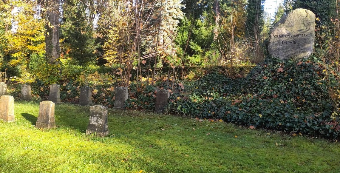 Alte Gräbsteine auf einer Rasenfläche, rechts im Hintergrund ein großer Stein mit der Aufschrift „Den tapferen Kriegern 1914–18“