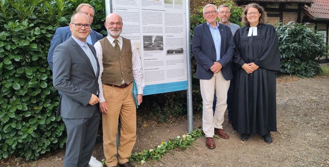 Eine Gruppe von fünf Männern und einer Frau, die Pfarrerin , stehen links und rechts der Tafel. Sie lächeln in die Kamera.