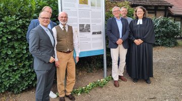 Eine Gruppe von fünf Männern und einer Frau, die Pfarrerin , stehen links und rechts der Tafel. Sie lächeln in die Kamera.