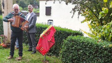 Links im Bild zwei Männer. Einer spielt Akkordeon. Hinter ihnen eine Stele, rechts von ihnen die verhüllte Pulttafel. Im Vordergrund das Grab.