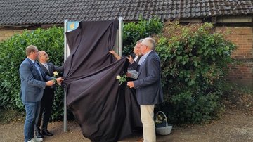 Mehrere Männer und die Pfarrerin halten ein schwarzes Tuch in den Händen und entfernen dieses von der noch weitesgehend verhüllten Tafel.