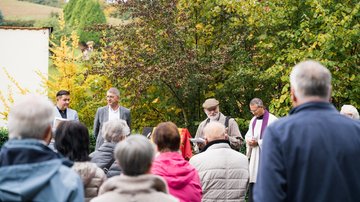 Eine Gruppe von Menschen, von hinten fotografiert. Dahinter vier Männer mit dem Gesicht zur Kamera. Darunter ein Pfarrer. 