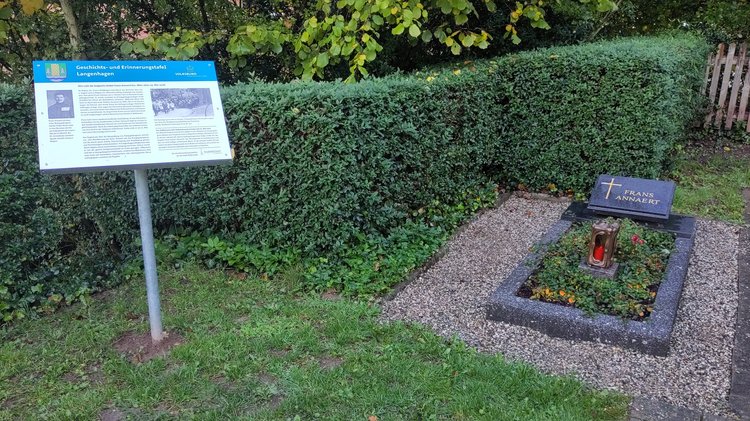 Rechts im Bild das gut gepflegte Grab. Links die neue Pulttafel mit Text und zwei Fotos. Im Hintergrund eine Hecke.