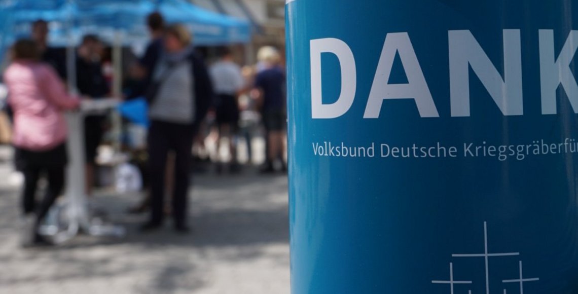 Im Vordergrund ein Ausschnitt einer blauen Volksbund Sammeldose. Angeschnitten ist darauf das Wort Danke zu sehen sowie die fünf Volksbundkreuze. Im Hintergrund, unscharf, eine Gruppe Menschen an einem Infostand des Volksbundes.