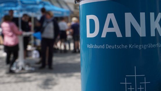 Im Vordergrund ein Ausschnitt einer blauen Volksbund Sammeldose. Angeschnitten ist darauf das Wort Danke zu sehen sowie die fünf Volksbundkreuze. Im Hintergrund, unscharf, eine Gruppe Menschen an einem Infostand des Volksbundes.