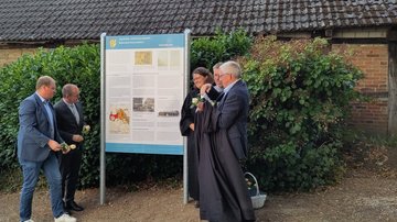 Die Tafel ist nun enthüllt. Die Personen betrachten sie, in den Händen halten einige jetzt weiße Rosen.