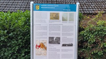 Das Bild zeigt den Busch vor dem nun die Tafel steht. Darauf Text in drei Spalten sowie diverse Abbildungen.