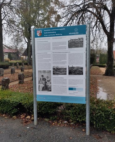 Die Tafel in einem Metallgestell vor einem Gräberfeld.
