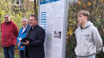 Ein Pfarrer steht vor der Tafel und spricht. Erhält eine kleine,  aufgeschlagene Mappe in der Hand. Links im Bild zwei Männer, rechts ein Schüler.